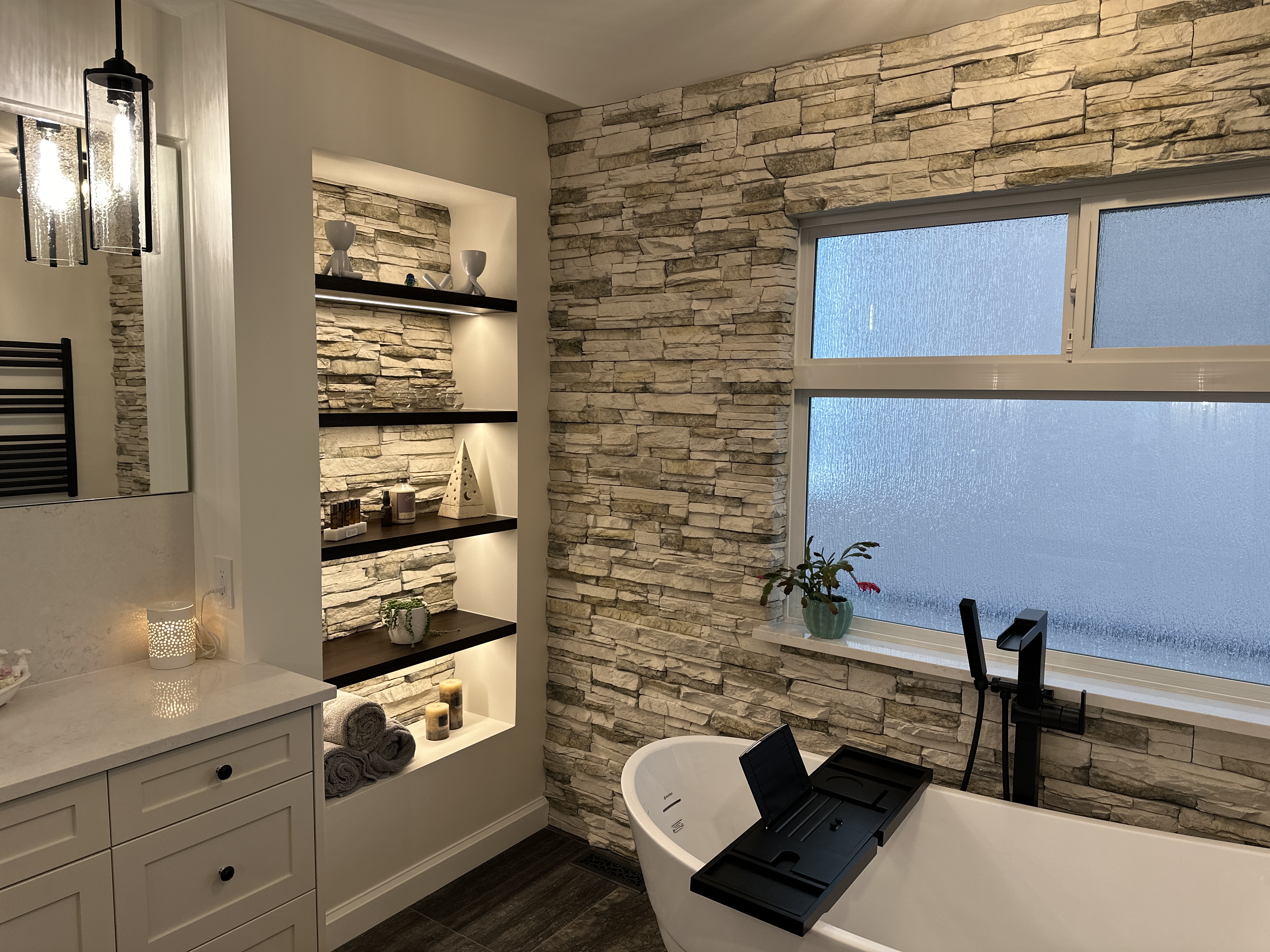Primary bathroom with a freestanding tub, black floor-mount filler, and a stacked-stone accent wall with lit shelving.
