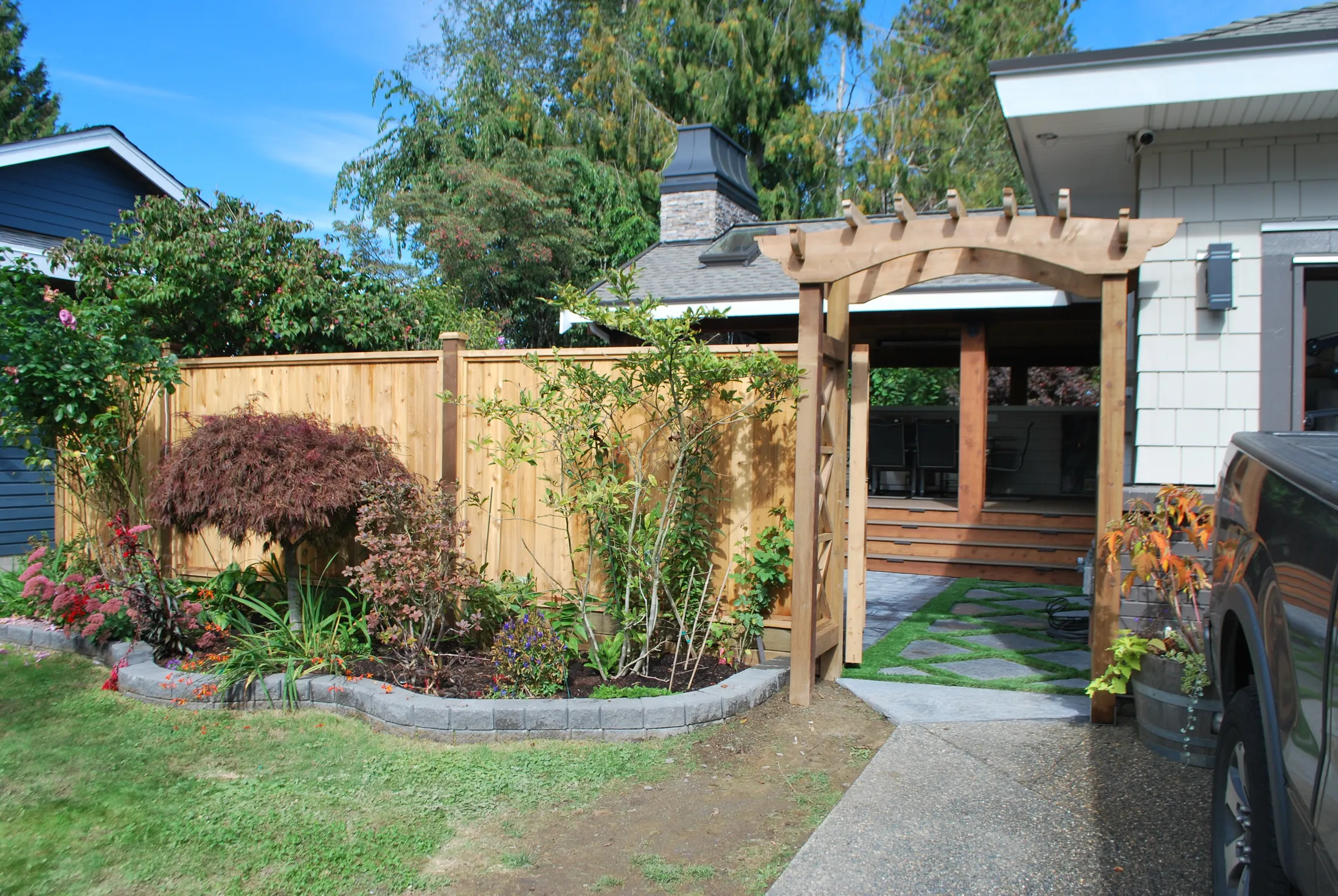 Backyard entrance with a cedar pergola gate, landscaped garden bed, and covered patio visible beyond the fence.