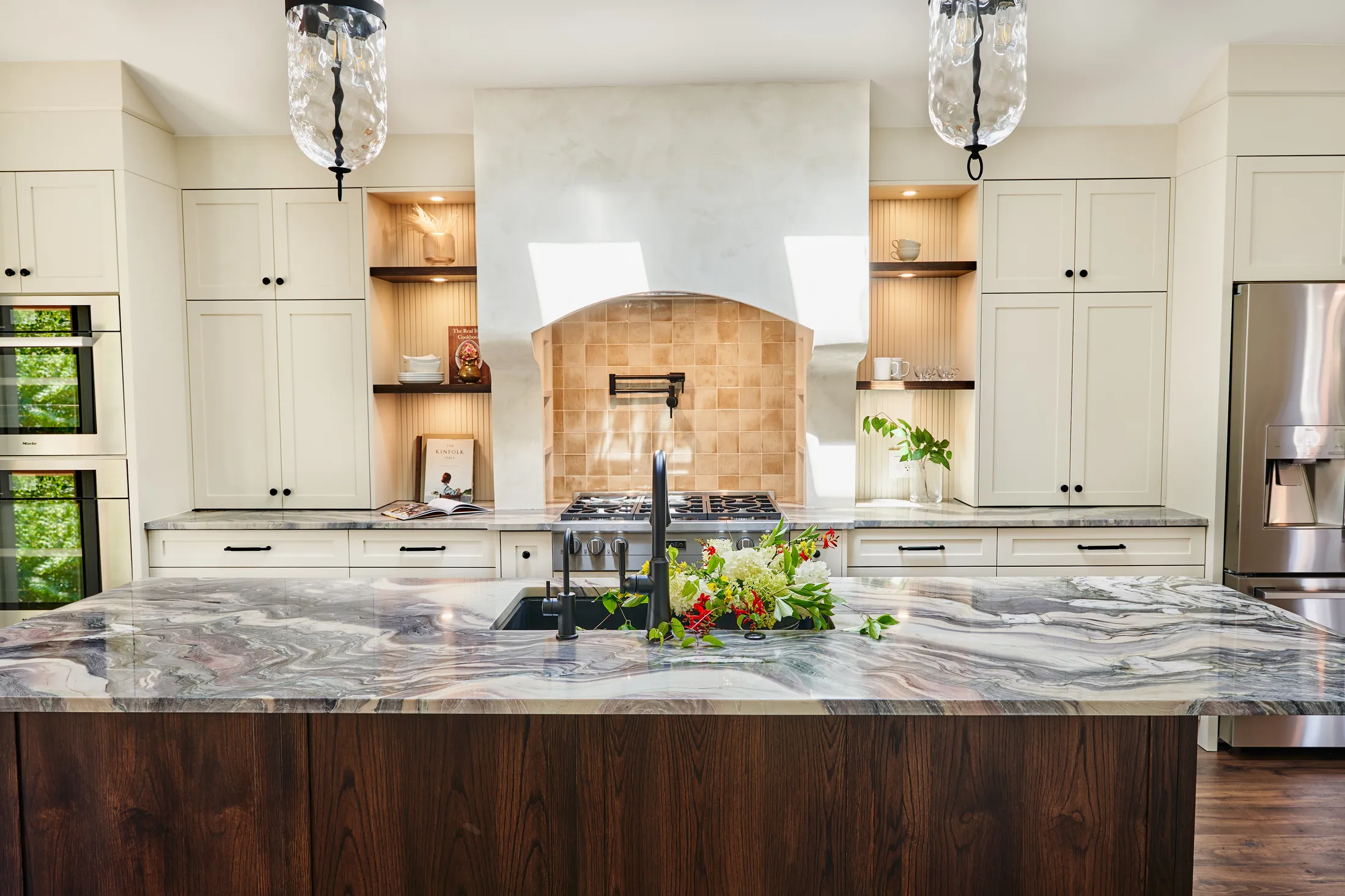 Kitchen island view toward the gas cooktop, arched plaster hood, tiled niche, and black pot filler.