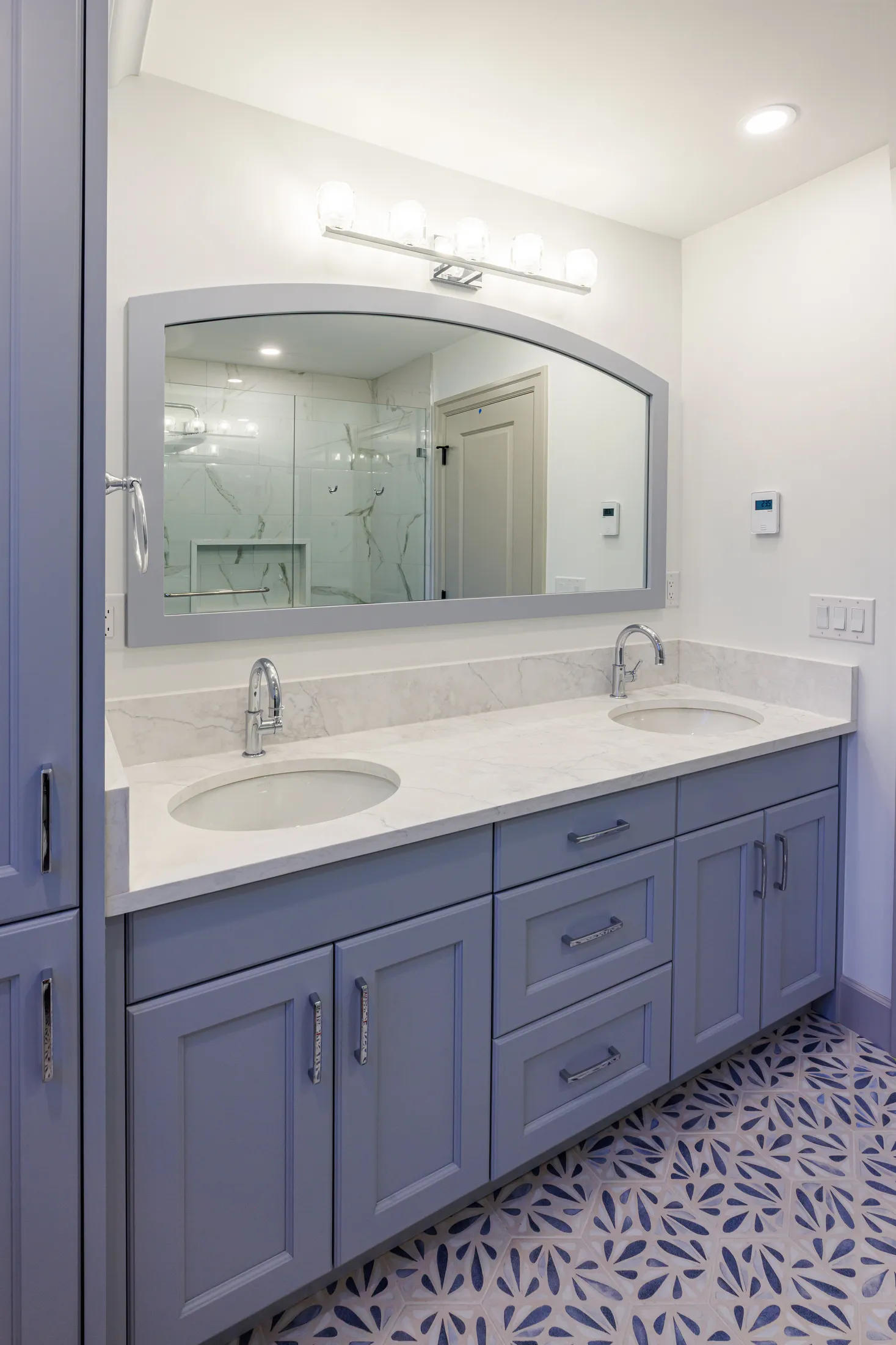 Primary bathroom double vanity in blue-gray cabinetry with marble counter and patterned tile floor.